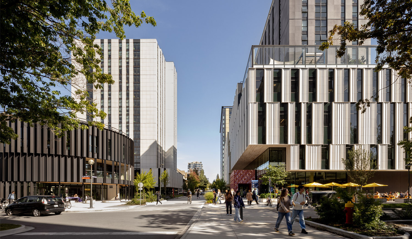 Wide view of a modern urban campus street with tall contemporary buildings on both sides, trees lining the sidewalks, and people walking and cycling under a clear blue sky.