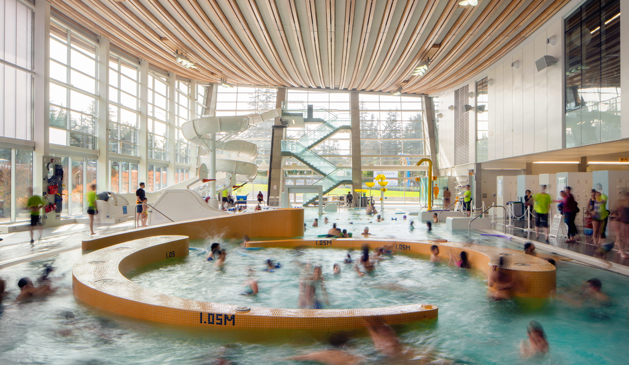People of all ages enjoying the leisure pool with its feature spiral water slide at the deep end of the pool. The high ceilings and ample nature light make the space an inviting place to be.