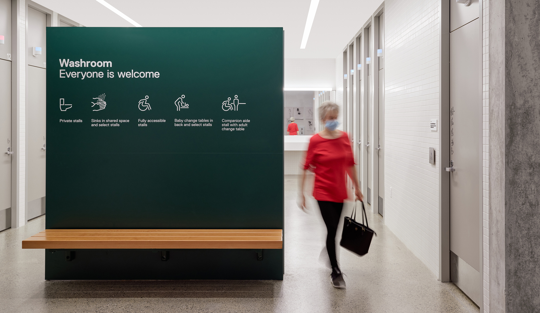 A lady walking by a sign that says "Washroom, Everyone is welcome" with a set of icons.