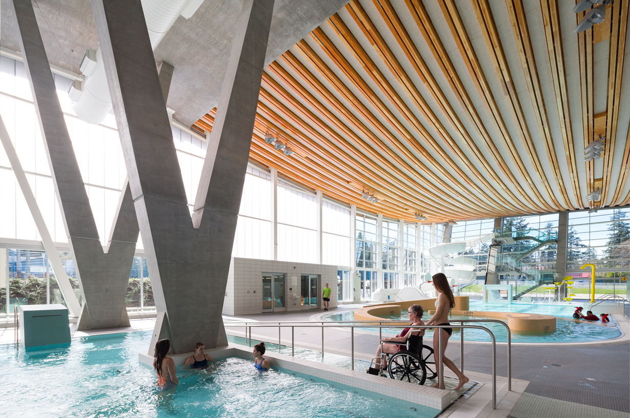 A girl pushing someone in a wheelchair into the indoor pool with high, wooden ceilings