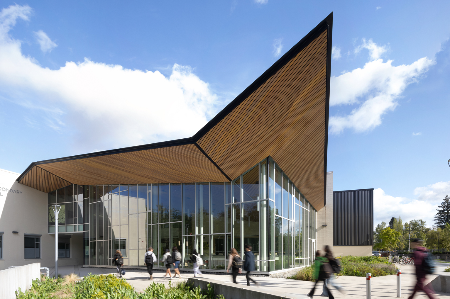 Students walking past the outside corner of R.E. Mountain Secondary School to go in. The corner of the building includes floor to ceiling windows and a wooden roof that protrudes up into a point.