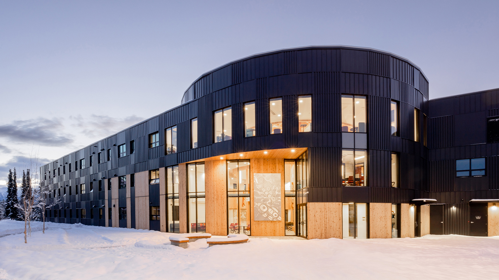 An outside view of Wii Gyemsiga Siwilaawksat, which is a long building that rounds at the entrance. The exterior has black panelling and wood, showcasing a large Indigenous art piece as you enter the building.