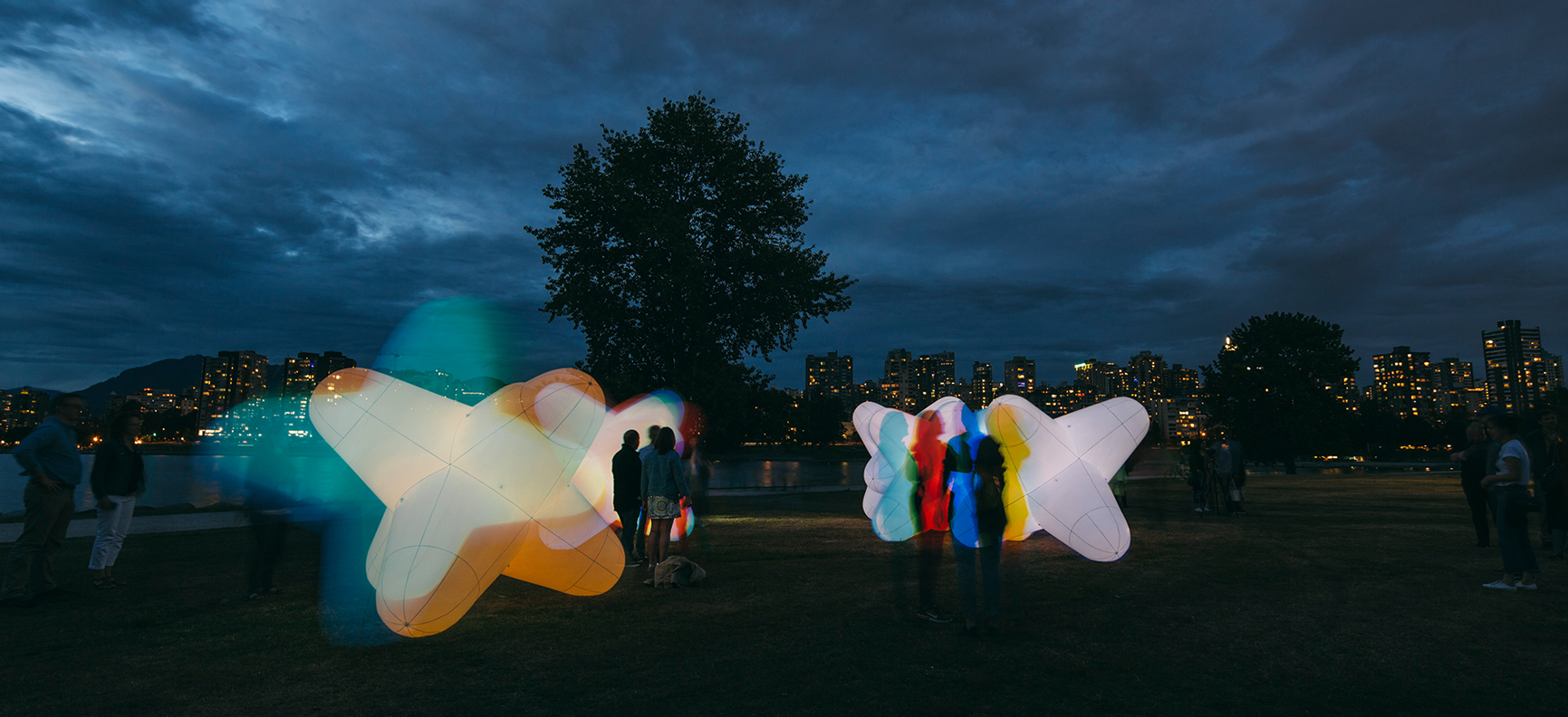 Night photo from a Jax activation. Various inflatable shapes, that appear similar to shapes from the game "Jacks" appear in various bright colours under a cloudy dark sky, with silhouettes of various people interacting with them.