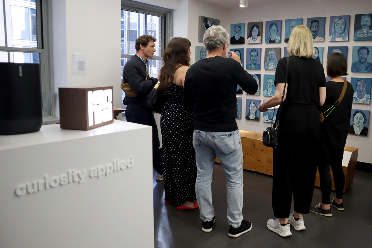 A group of people mingle while looking at portraits of various hcma-ers, created during Kyla Bourgh's residency, Comforting Illusions. To the left, the office front desk is in view, which has subtle white lettering that reads, "curiosity applied".