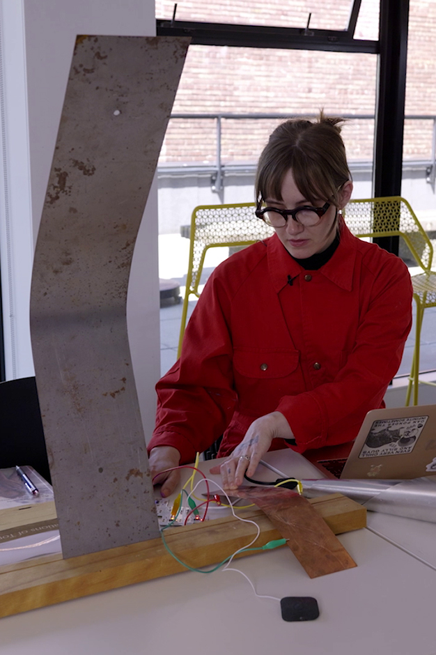 Edie with a large metal sculpture they created. Viewers can create a reverberant sound composition via body conductivity.