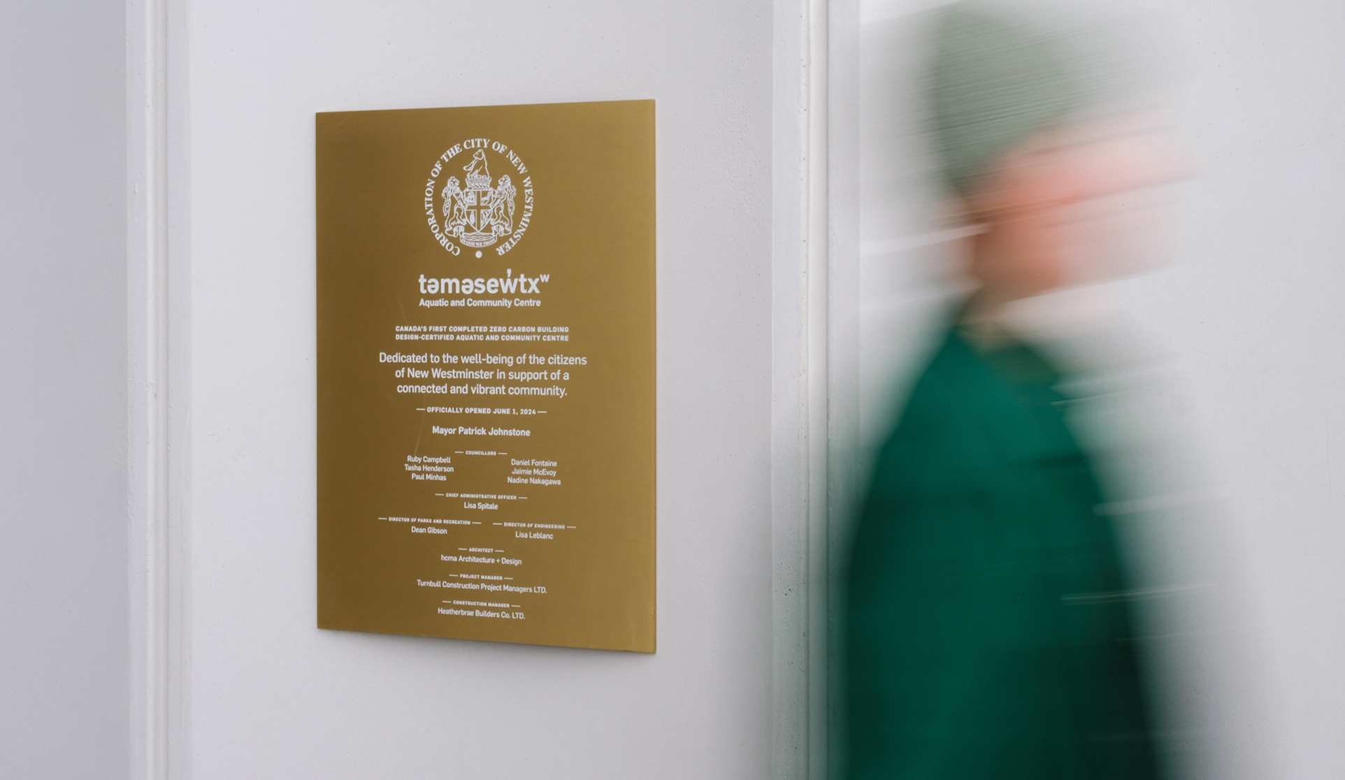 A blurred person walks past a gold plaque mounted on a white wall. The plaque reads "tam̓əsəw̓txʷ Aquatic and Community Centre" and commemorates the official opening of Canada's first completed zero-carbon building design-certified aquatic and community centre in New Westminster on June 8, 2024. The plaque includes names of project contributors, including the mayor, city council members, design and construction teams.