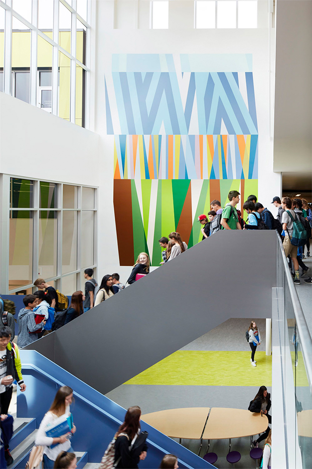 Students walk up and down a wide staircase within the bright, modern Royal Bay Secondary School atrium, with large windows and a colourful abstract mural on the wall behind them.