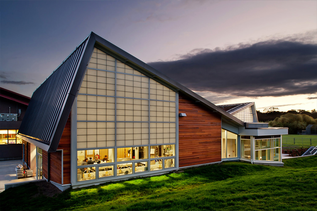 Evening view of the Arts Centre at Cedar Hill, a gabled building with translucent wall panels glowing from interior light, wood siding, and a sloped green lawn in the foreground. Through the windows, people are working away in the studio.