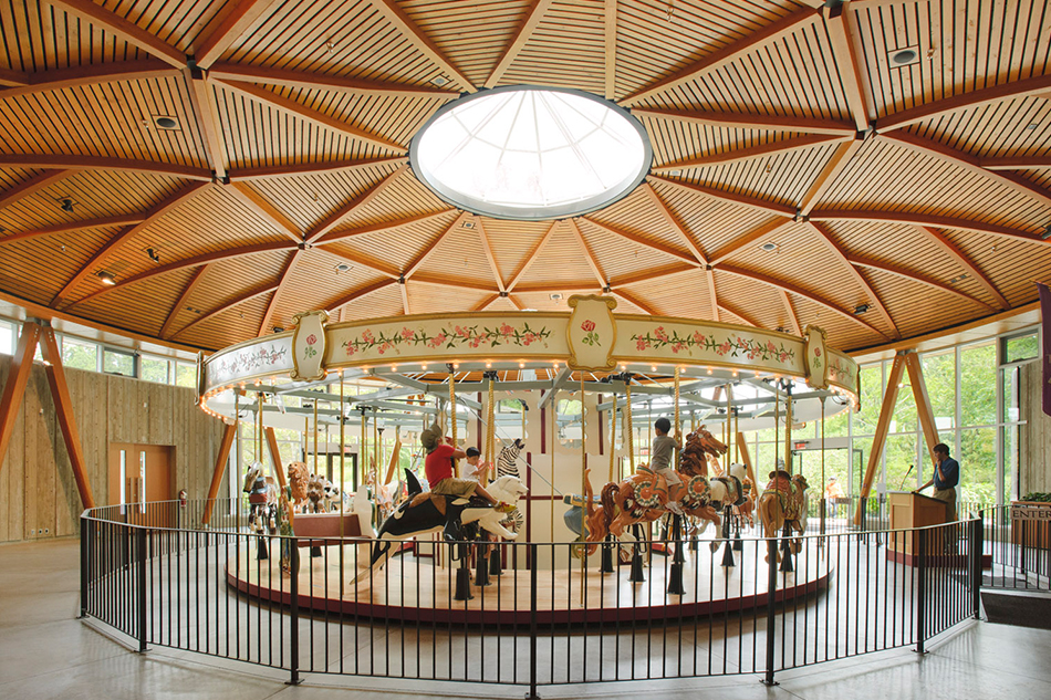 Children ride horses on a traditional carousel, housed inside the wood-framed Butchart Gardens Children's Pavilion, with a geometric timber ceiling and circular skylight.