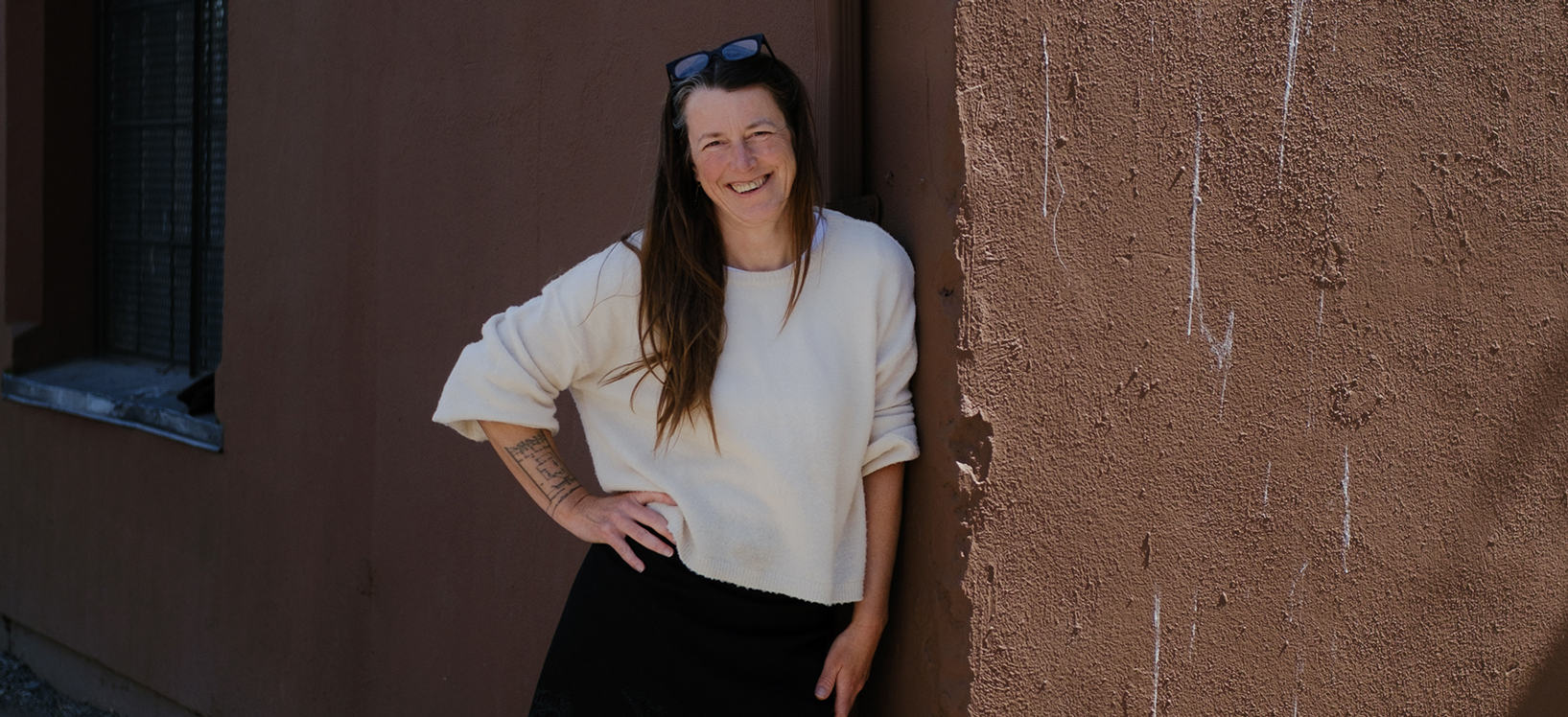 Photo of Heidi Nesbitt. She leans against a textured brown wall, smiling at the camera.